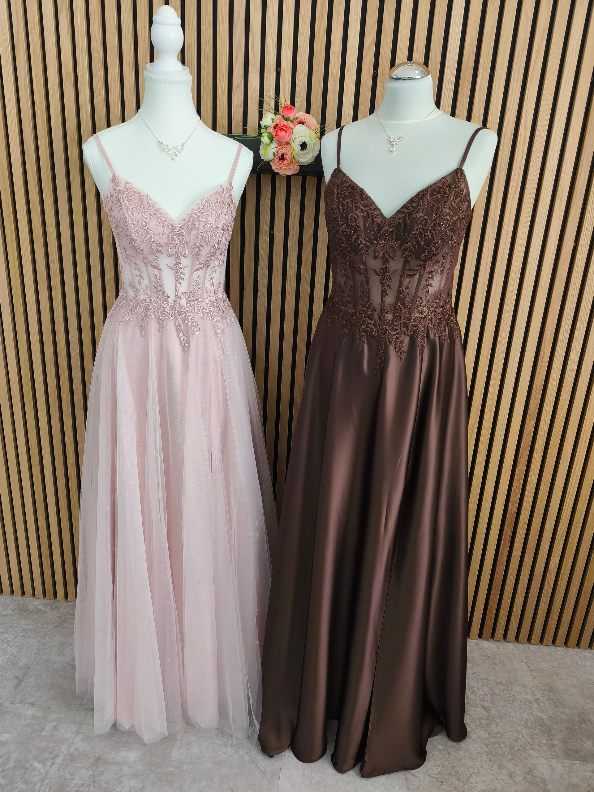 Zwei elegante Kleider an Schaufensterpuppen: eines in zartem Rosa, eines in Dunkelbraun, vor einer Wand aus Holzlatten.