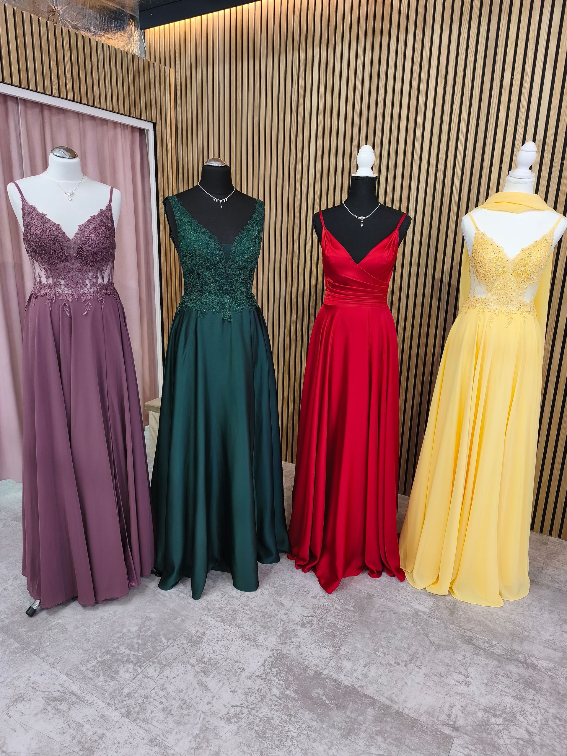Vier elegante Abendkleider an Schaufensterpuppen: malvenfarben, grün, rot und gelb vor einer Wand aus Holzlatten.