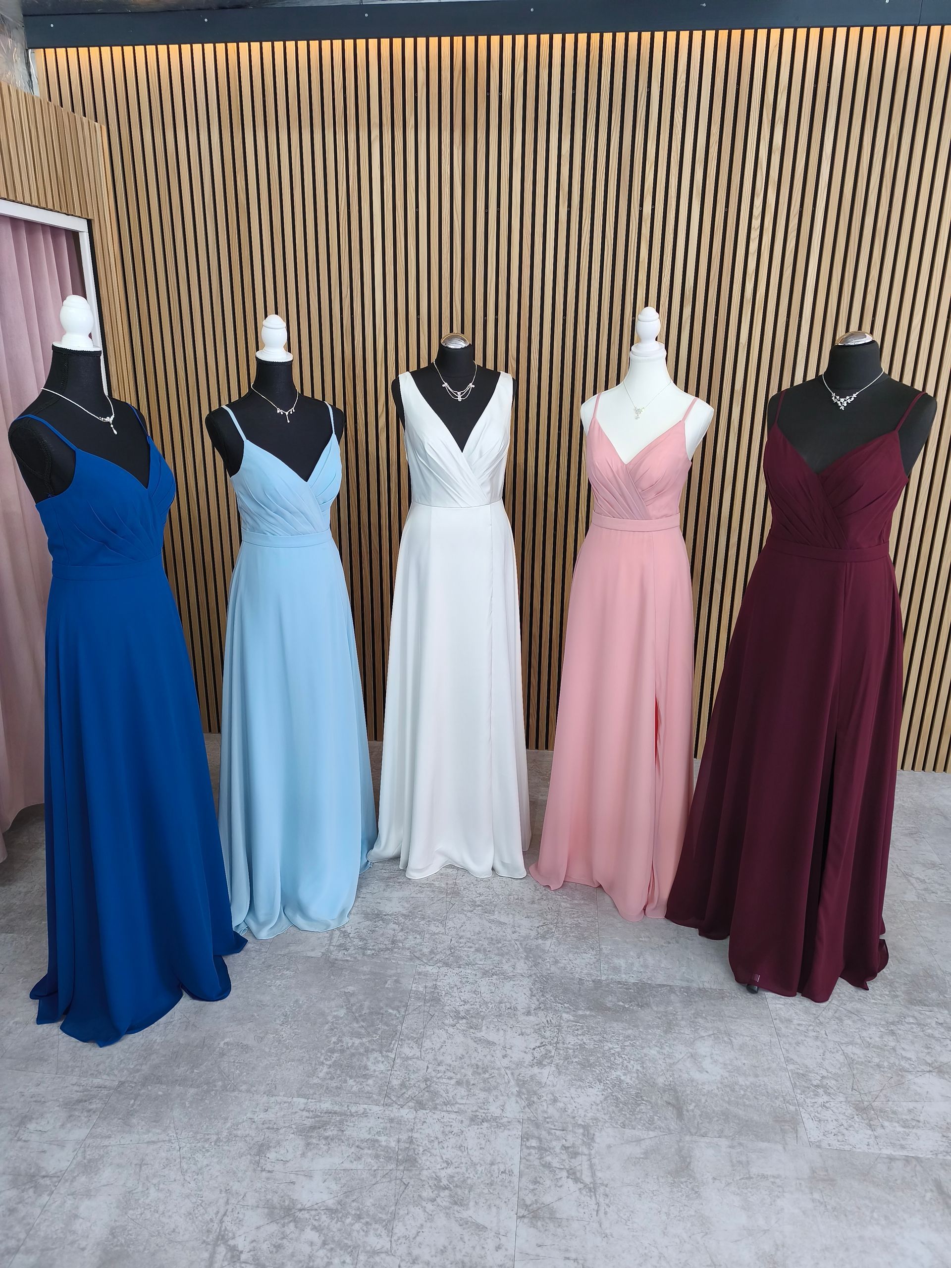 Fünf Schaufensterpuppen in langen, formellen Kleidern in Blau, Hellblau, Weiß, Rosa und Burgunderrot sind im Innenbereich ausgestellt.
