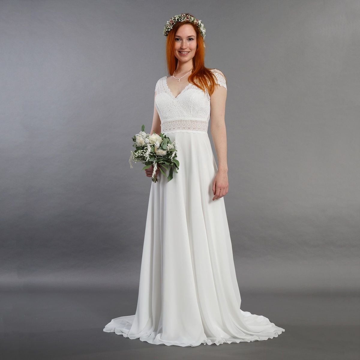 Frau in einem weißen Kleid hält einen Blumenstrauß vor grauem Studiohintergrund