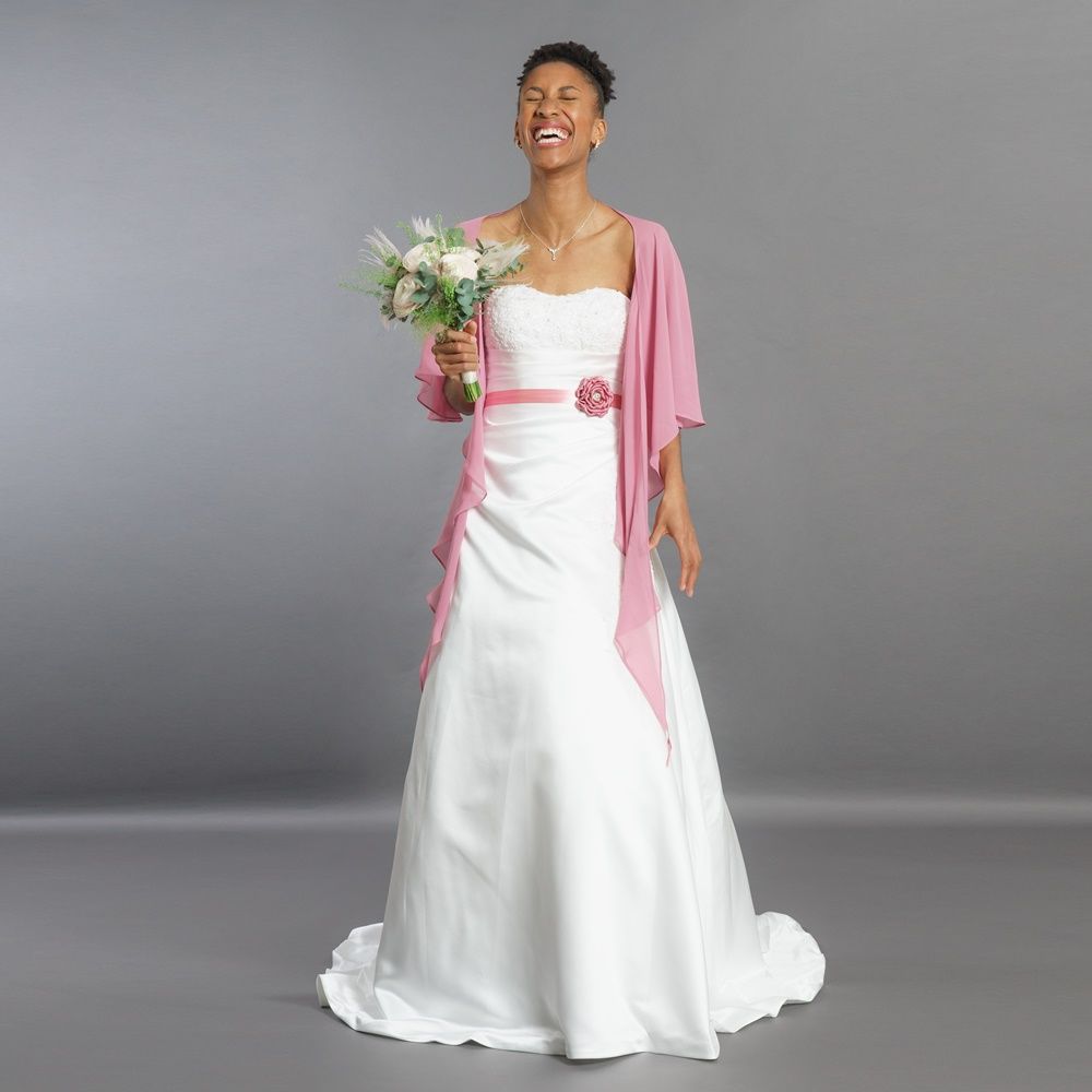 Frau in weißem Kleid und rosa Schal hält einen Blumenstrauß vor grauem Studiohintergrund
