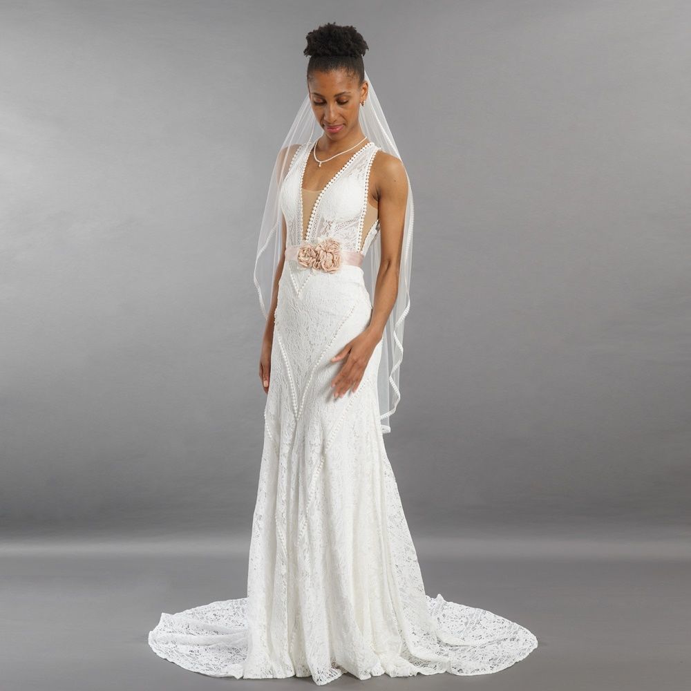 Eine Braut in einem weißen Spitzenkleid und Schleier steht vor einem grauen Studiohintergrund und blickt nach unten.