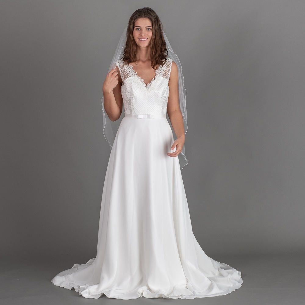 Eine Braut in einem weißen Hochzeitskleid mit langem Schleier steht vor einem grauen Hintergrund.