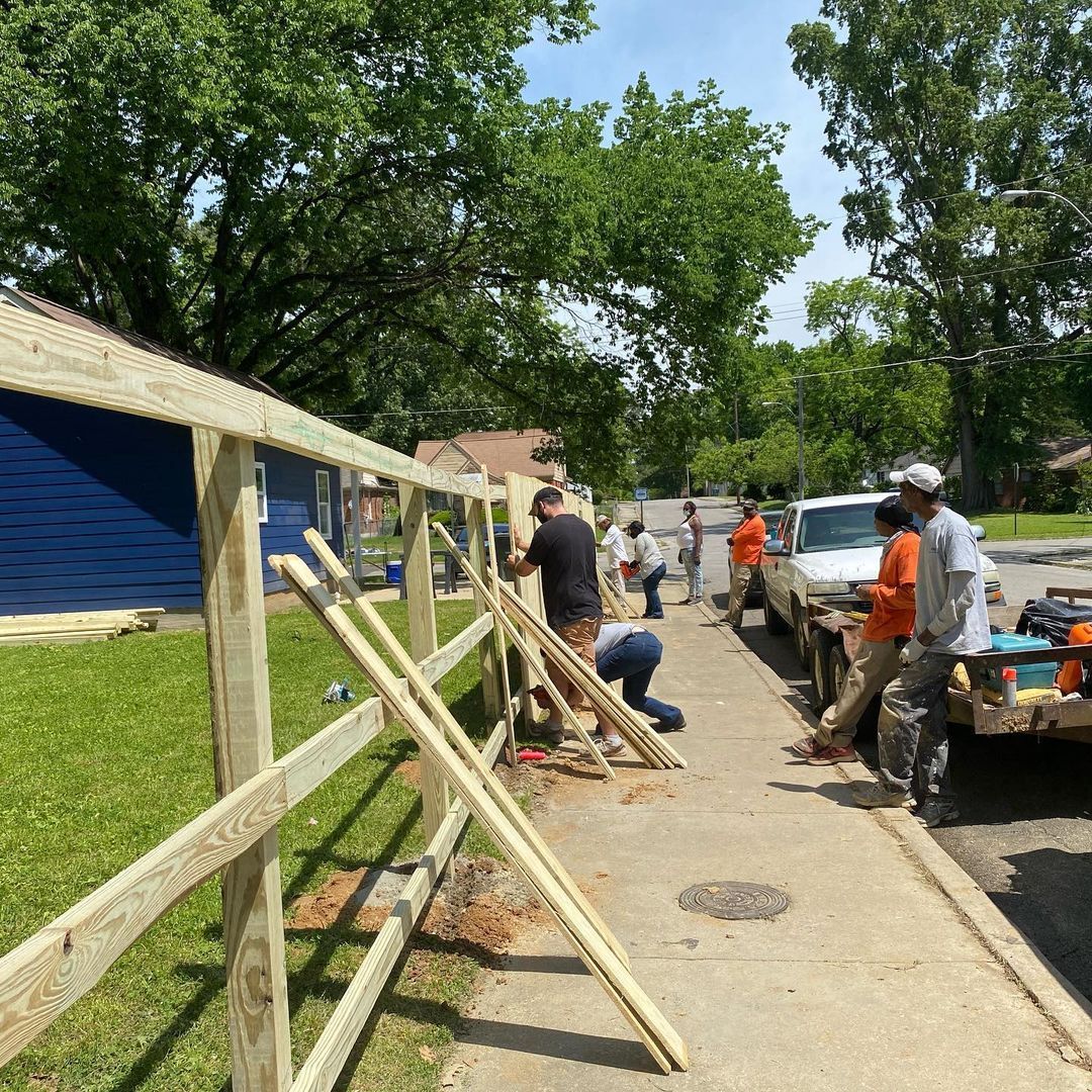 Workers repairing a wooden fence along a tree-lined sidewalk beside parked vehicles.