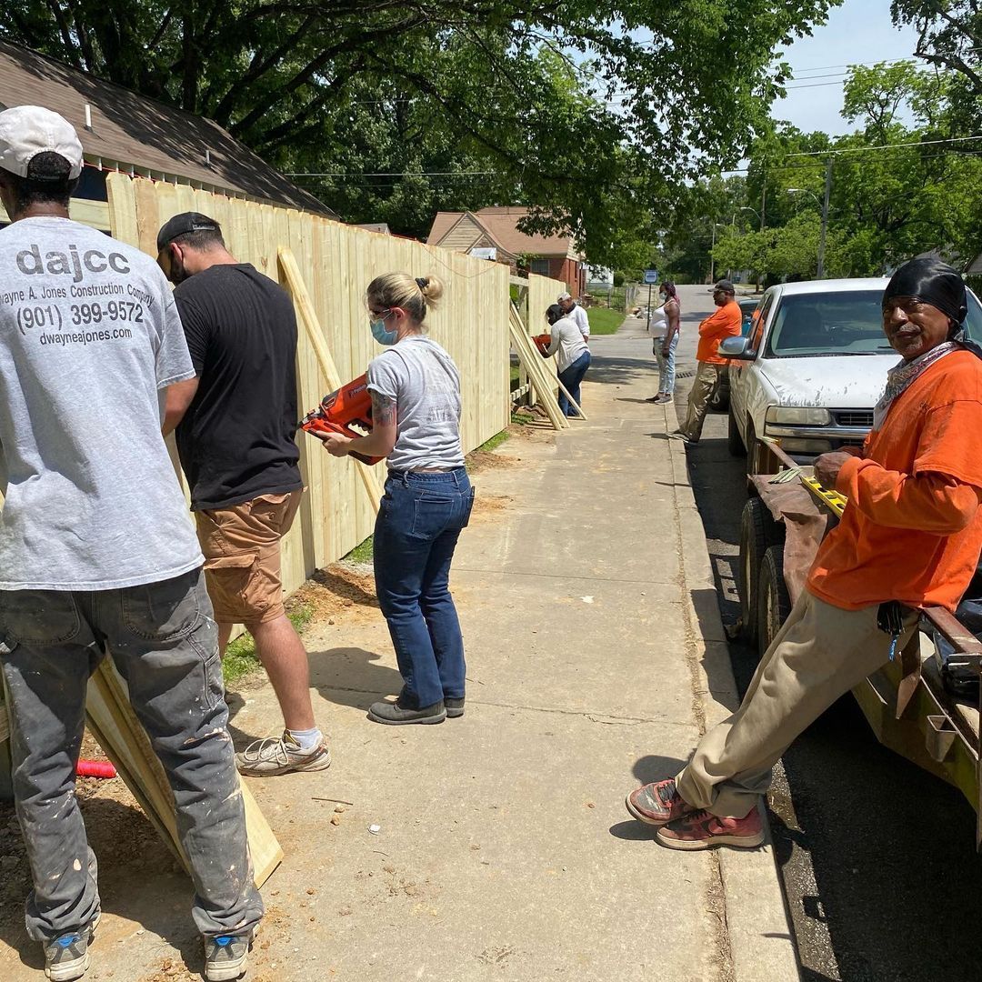 People standing and working along a sidewalk beside parked cars and a fence on a sunny day