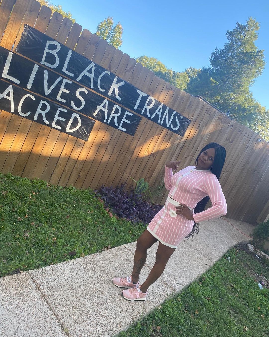 Person in a pink outfit stands on a sidewalk beside a fence painted “Black trans lives are sacred.”