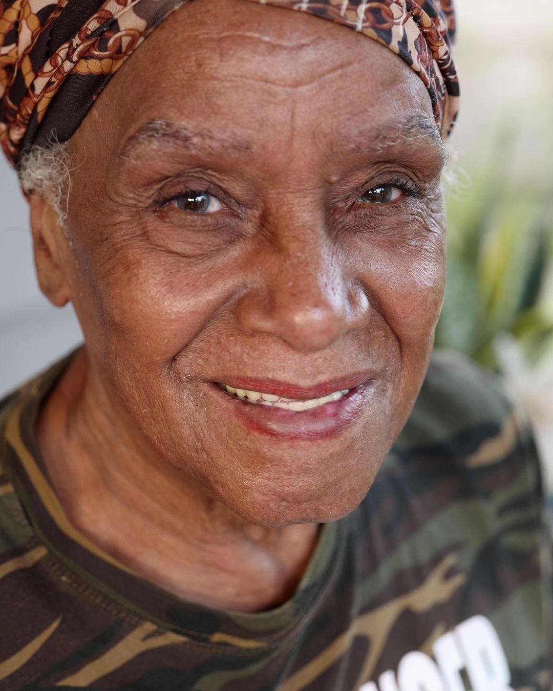 Close-up of a smiling person wearing a patterned headscarf and camouflage shirt outdoors.
