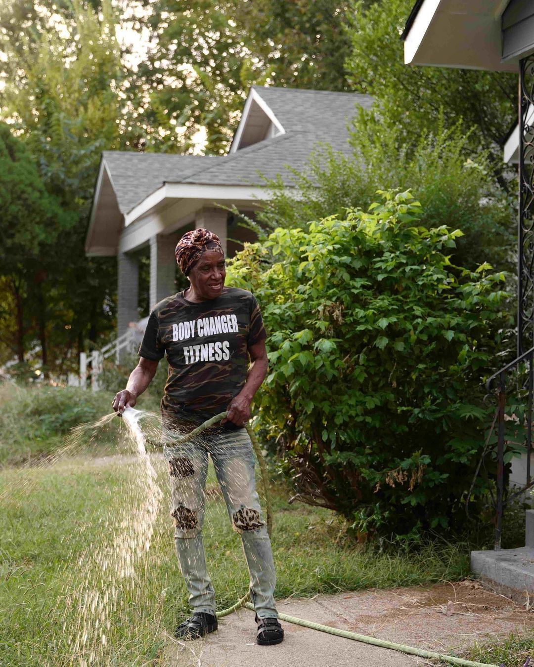 Person spraying water on a sidewalk beside a green hedge and house.