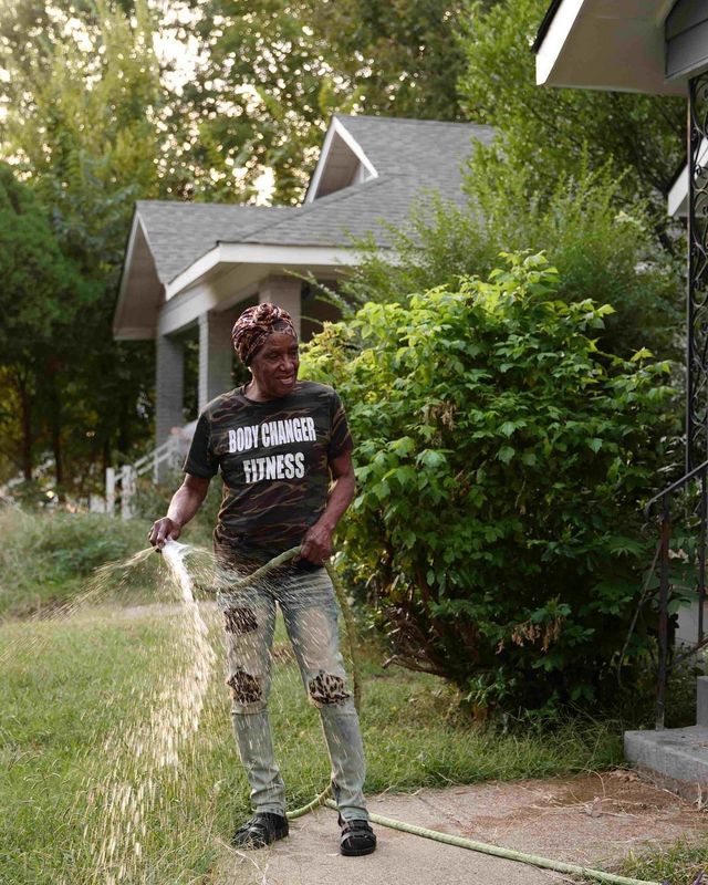 Person spraying water in a driveway beside a house and green shrubs.