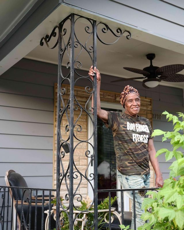 Person standing on a porch beside a wrought-iron post, smiling and holding it, with a ceiling fan overhead.