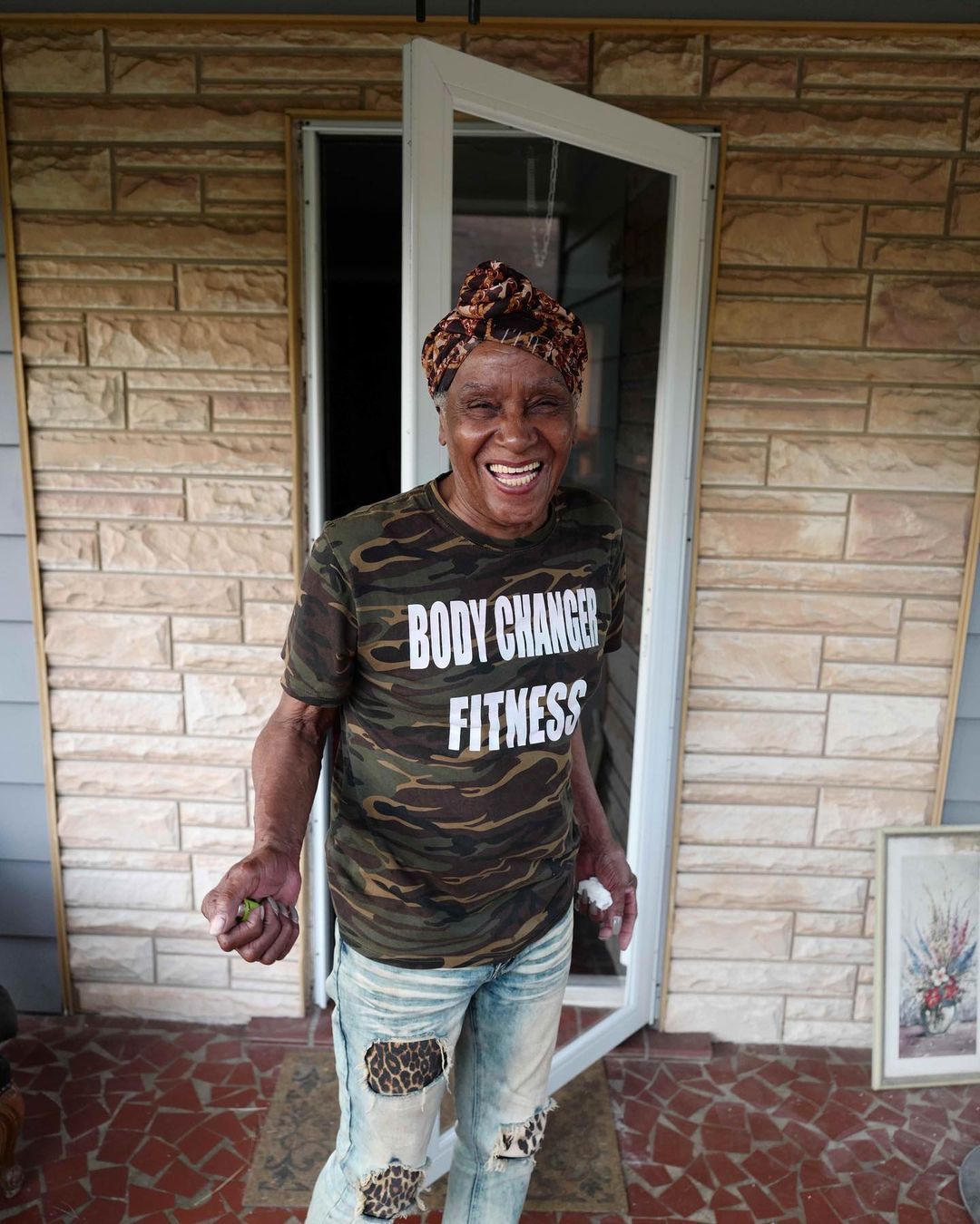 Smiling person in camo shirt and ripped jeans stands in a doorway on a brick porch