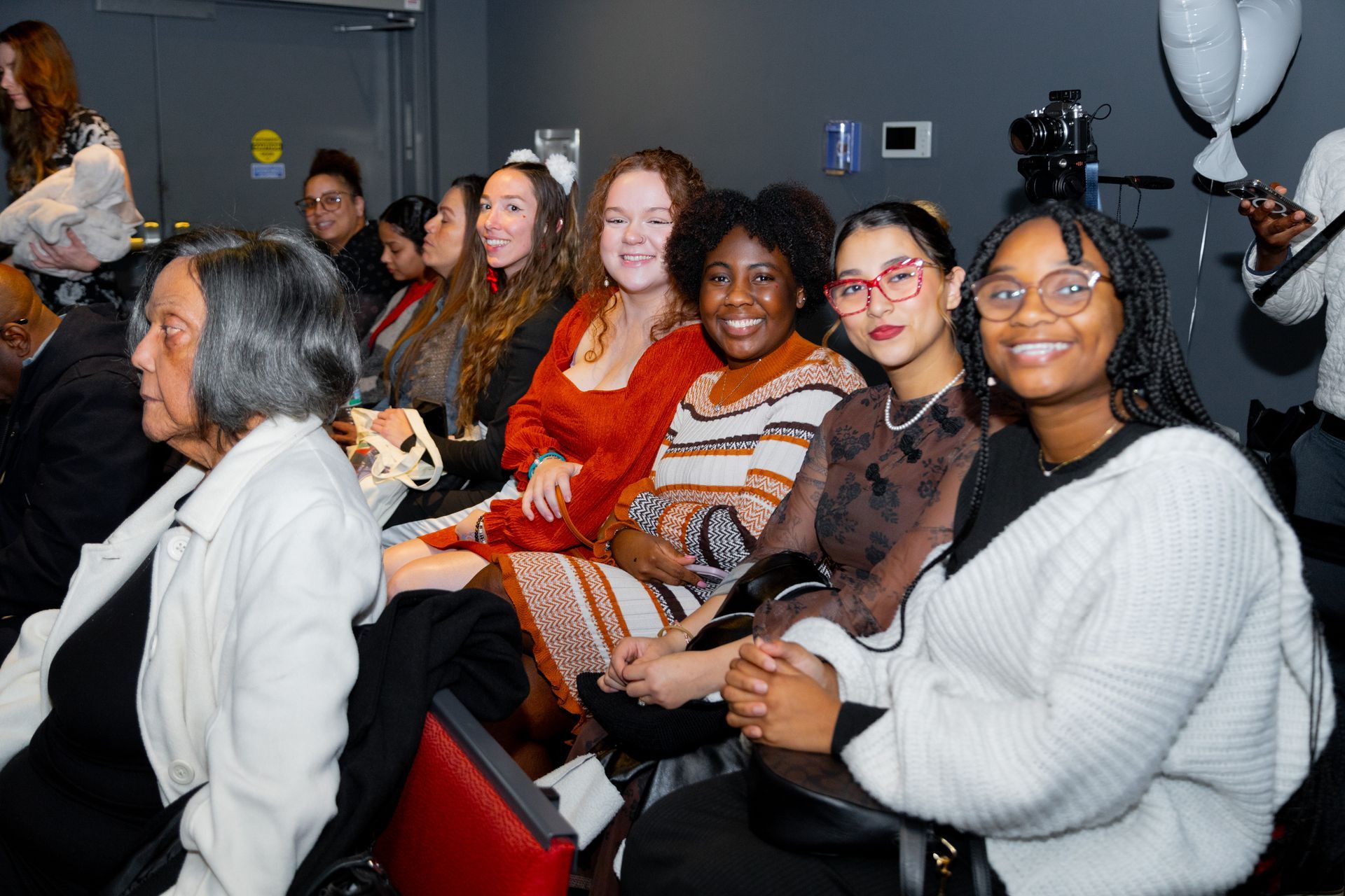 Seated audience smiling and chatting at an indoor event, with red chairs and dark walls.