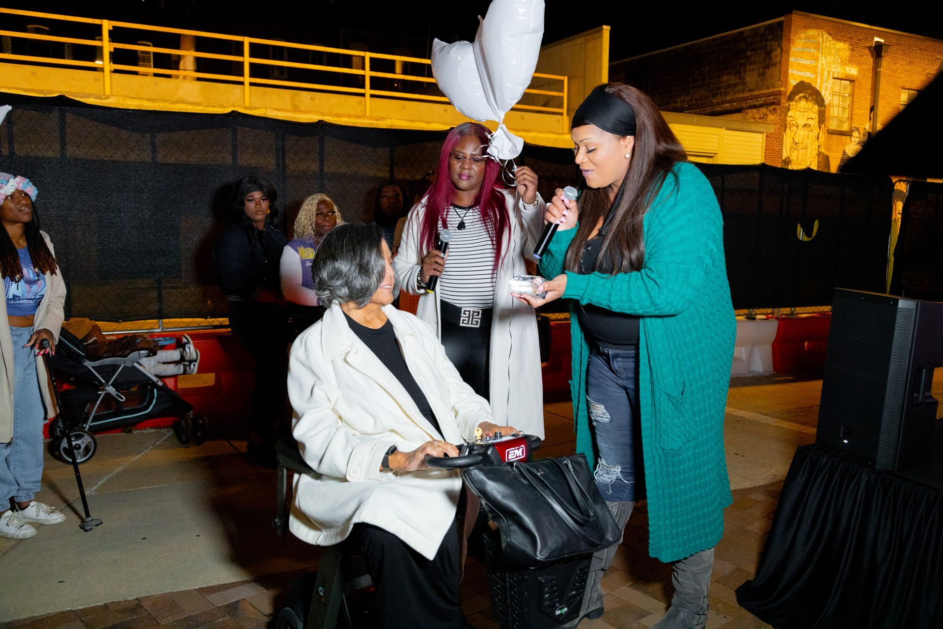 Woman in teal coat speaking to seated woman at a nighttime outdoor event with balloons and a crowd