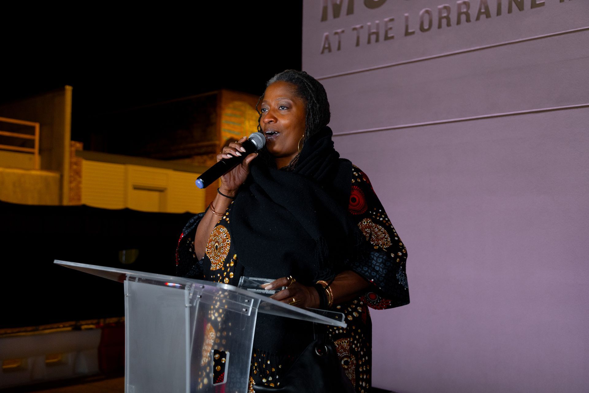 Woman speaking at a podium with a microphone during an event, wearing a black outfit with sparkling sleeves.