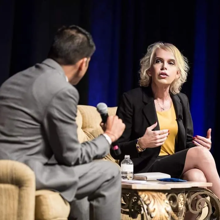 Two speakers onstage in a panel discussion, one holding a microphone, seated in armchairs