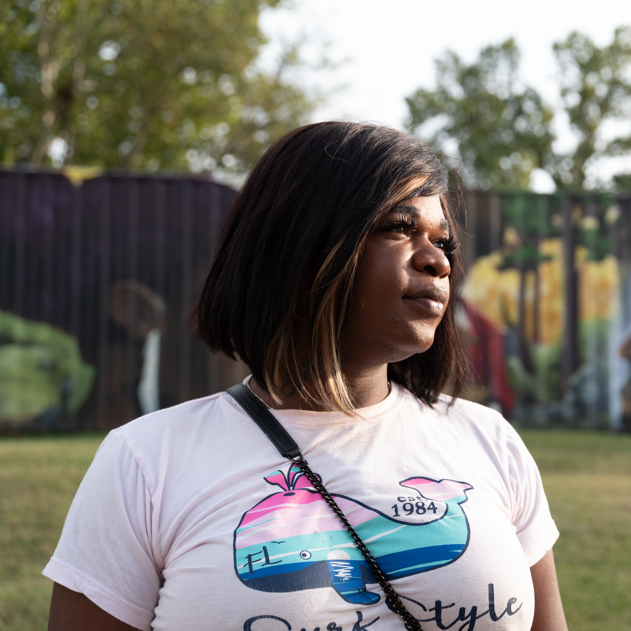 Woman in a white graphic T-shirt stands outdoors, looking right, with a mural and fence behind her.