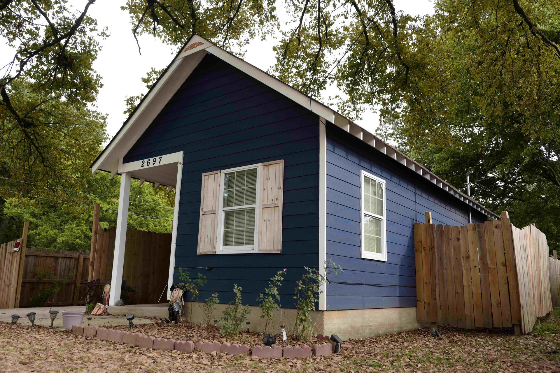 Small blue house with white trim, porch, and wooden fence in a leafy yard