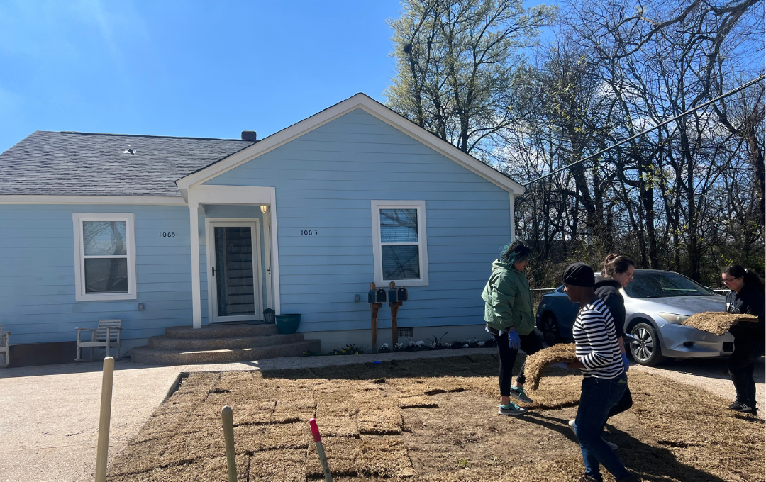 Small blue house with a front porch; two people stand in the yard near a parked car.