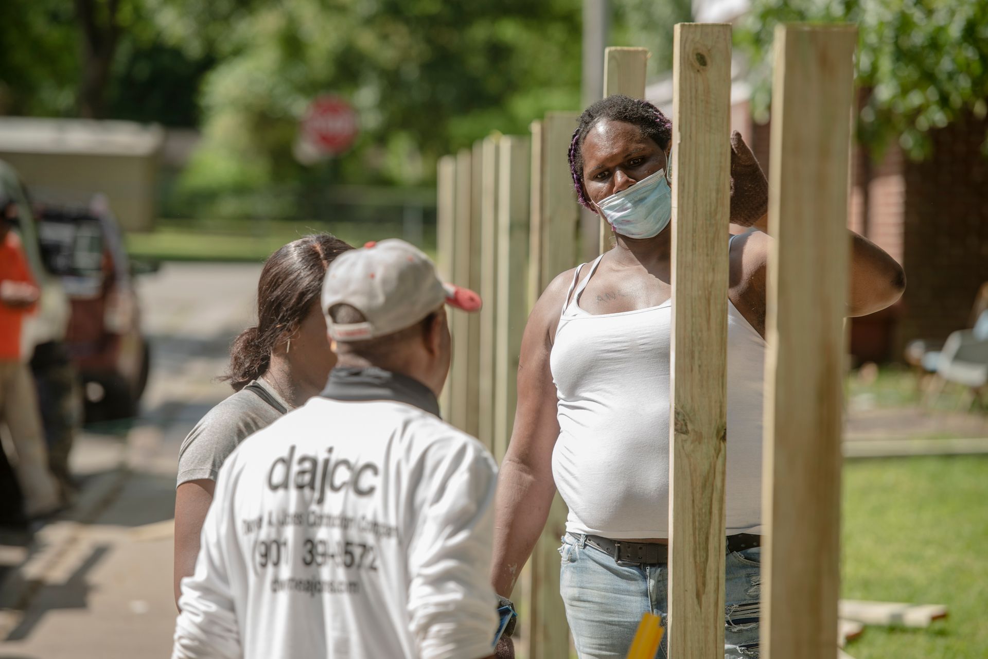 People talking outdoors near wooden posts, one wearing a white mask and tank top.