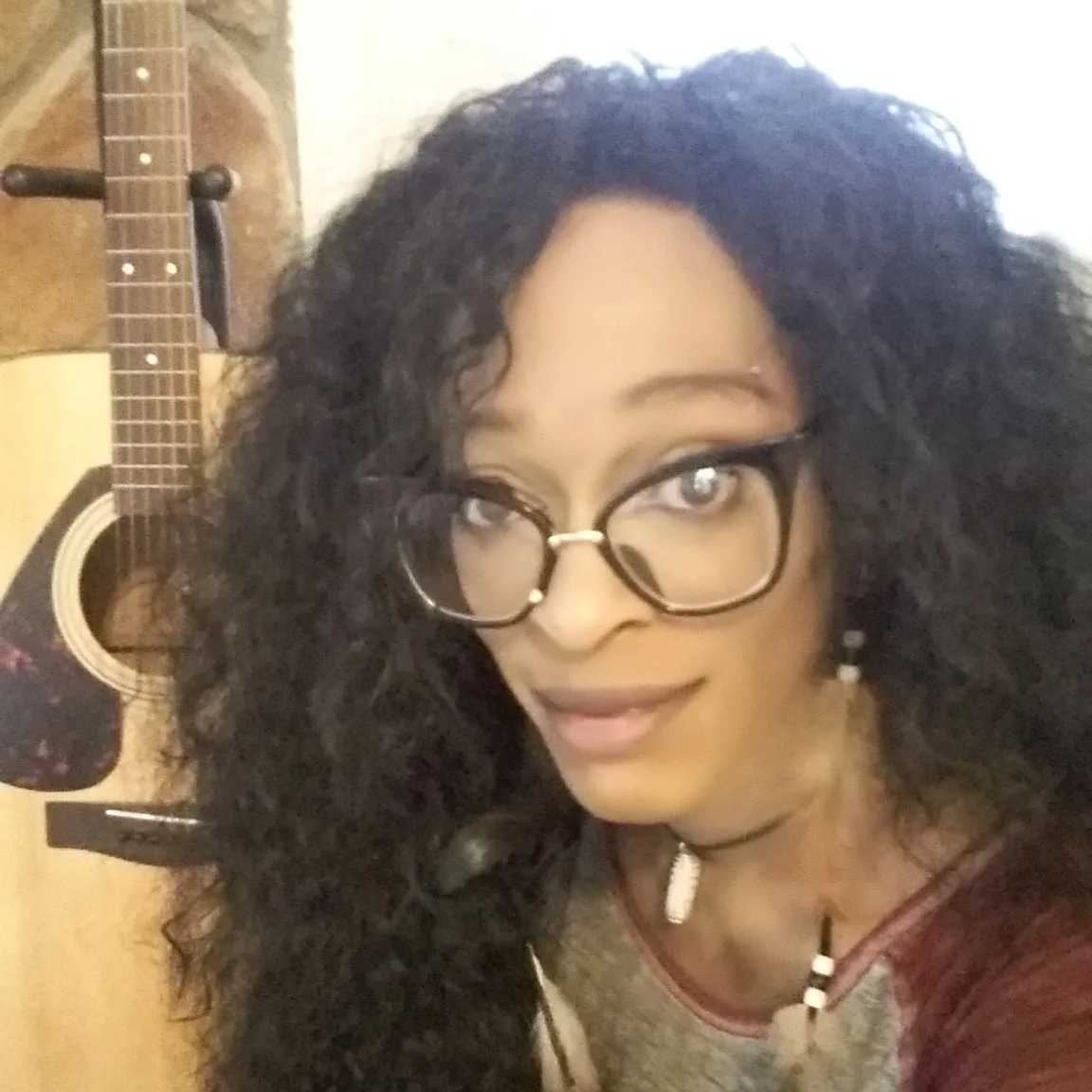 Selfie of a person with glasses and curly hair, holding a guitar in the background.