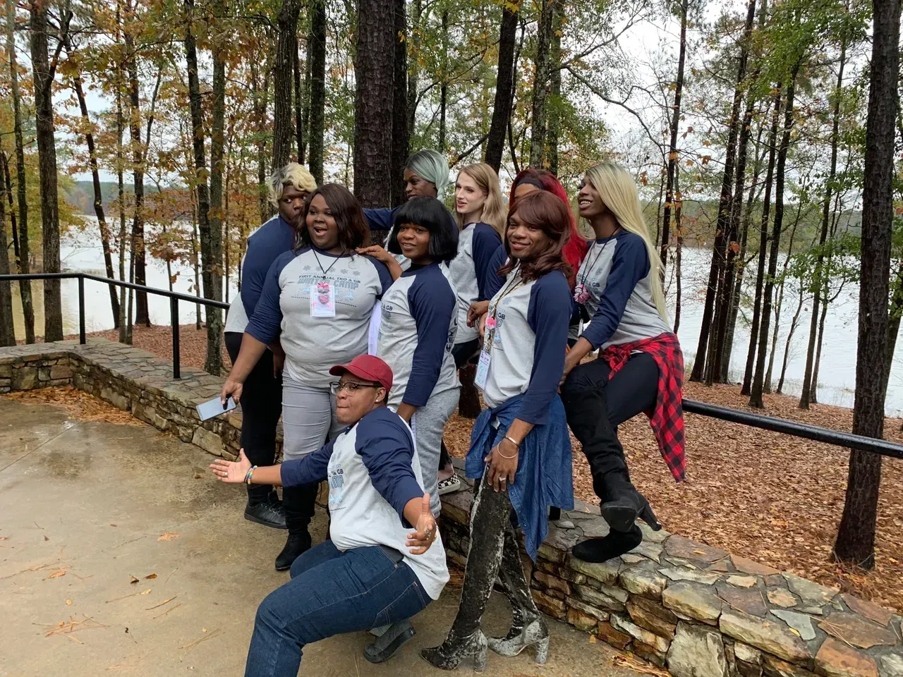 Group posing together outdoors by a stone wall near a lake and trees, some smiling and crouching.