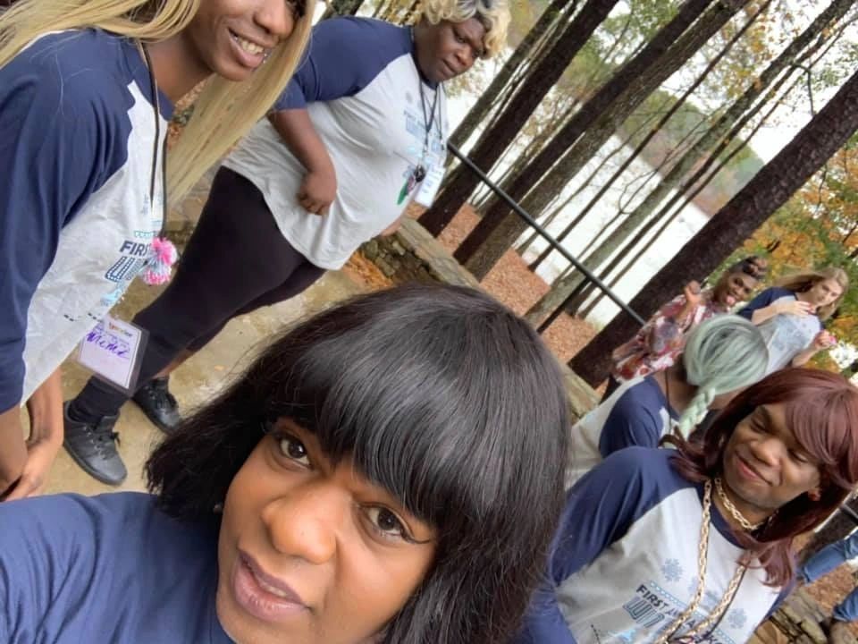 People standing outdoors in a wooded area, gathered closely and smiling at the camera.