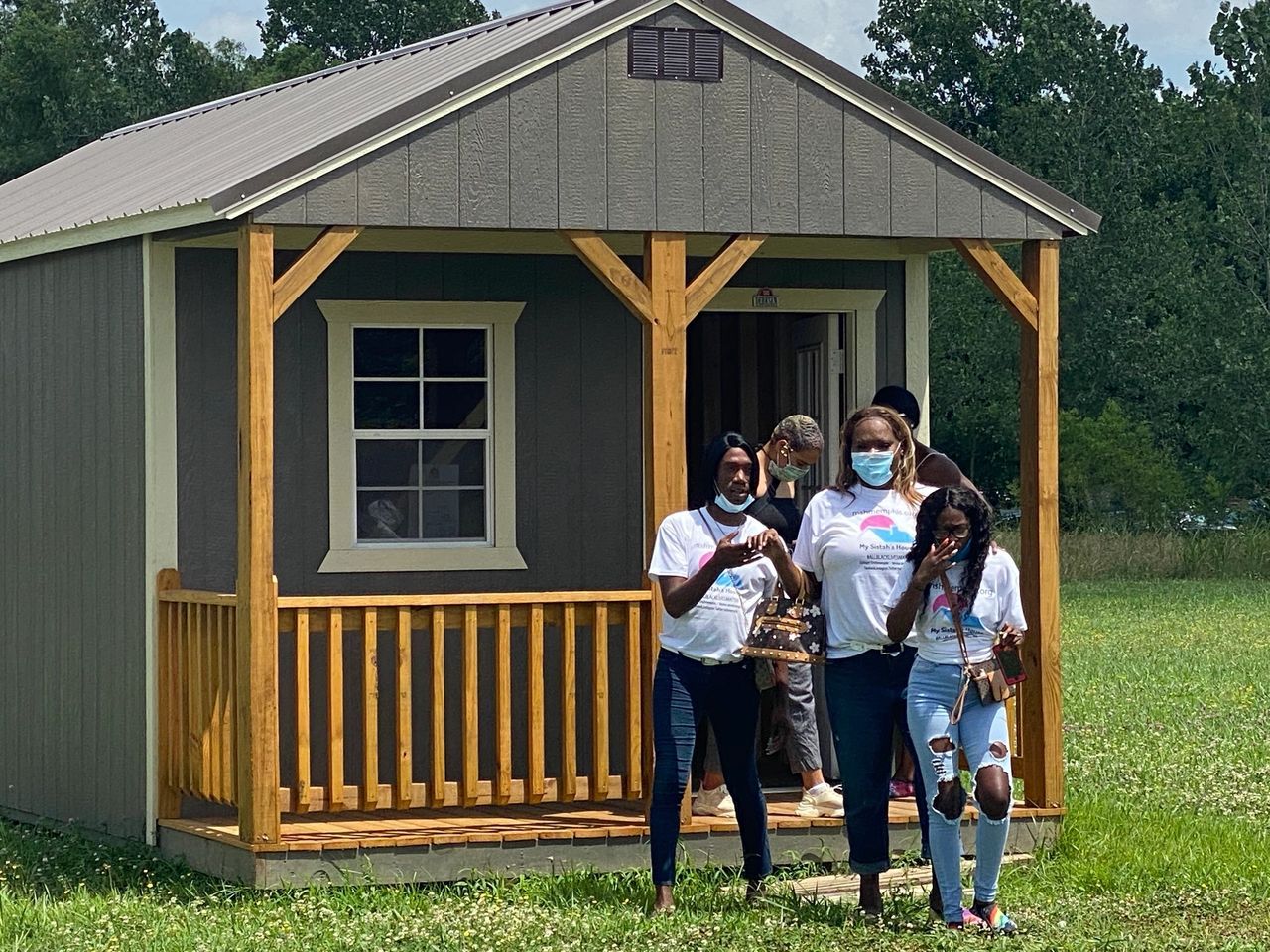 Three people stand in front of a small gray cabin with a wooden porch in a grassy field.