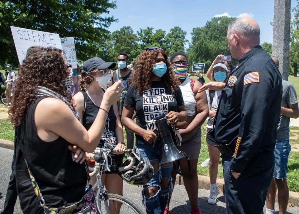 People gathered outdoors with bicycles, some wearing masks, in conversation near a roadside sign.