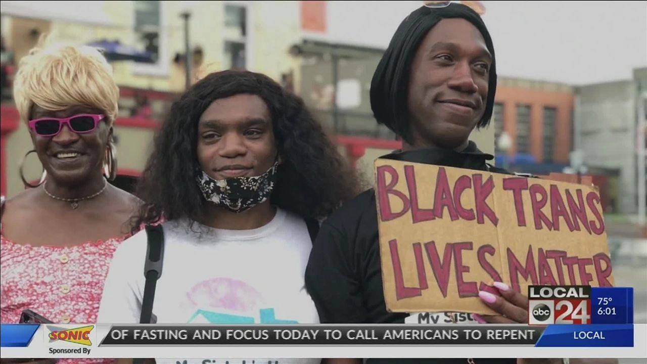 Three people at a protest, holding a “BLACK TRANS LIVES MATTER” sign, with a TV news banner below.