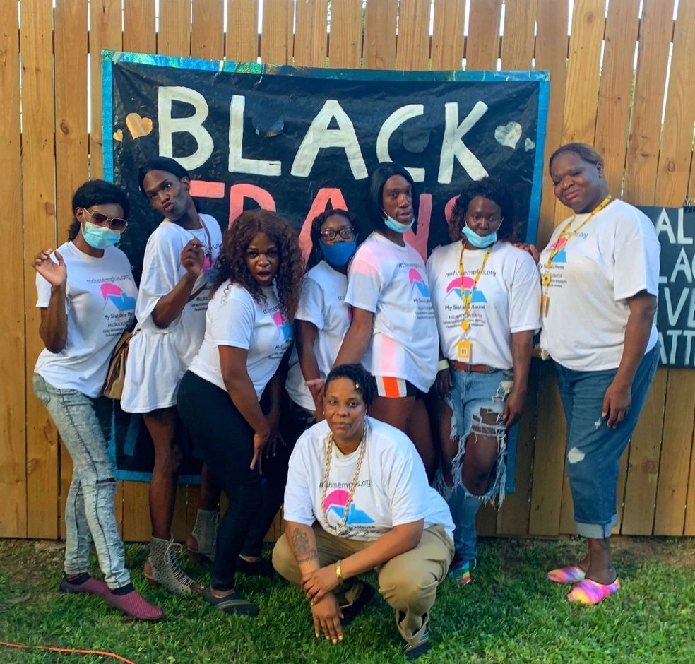 Group in white shirts posing outdoors in front of a “BLACK” banner at a wooden fence.
