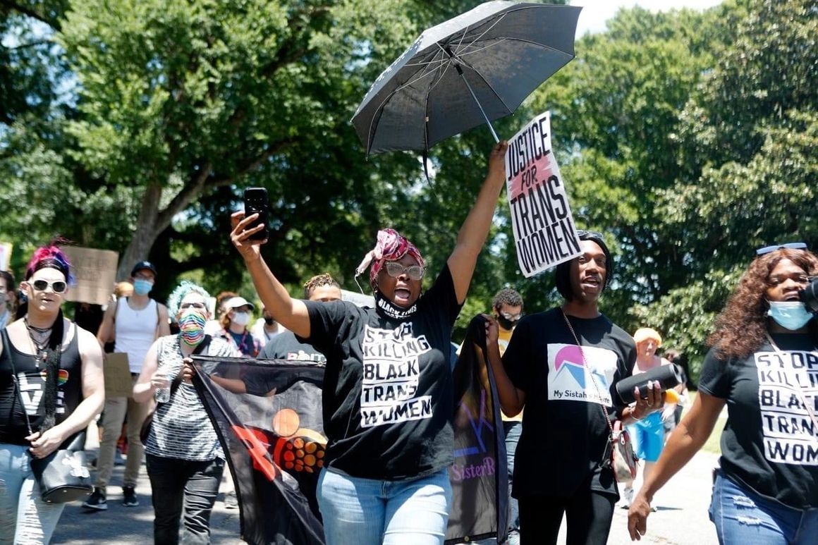 Protesters marching outdoors with signs, masks, and a black umbrella in a sunny street demonstration