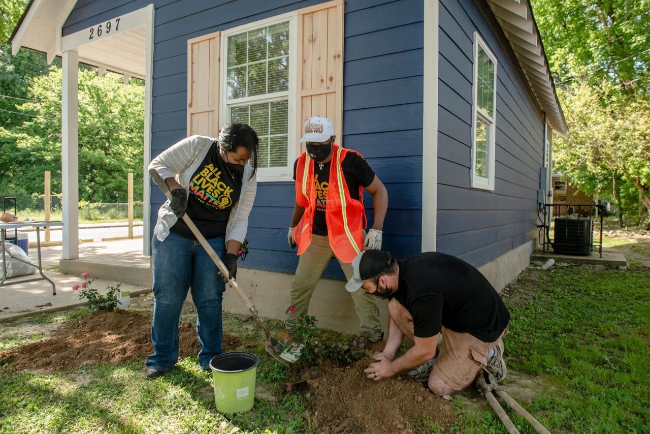 Three volunteers plant flowers beside a small blue house on a sunny day.