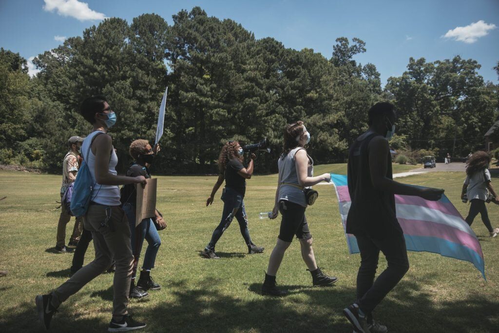 People walking across a grassy park holding a pink, blue, and white pride flag