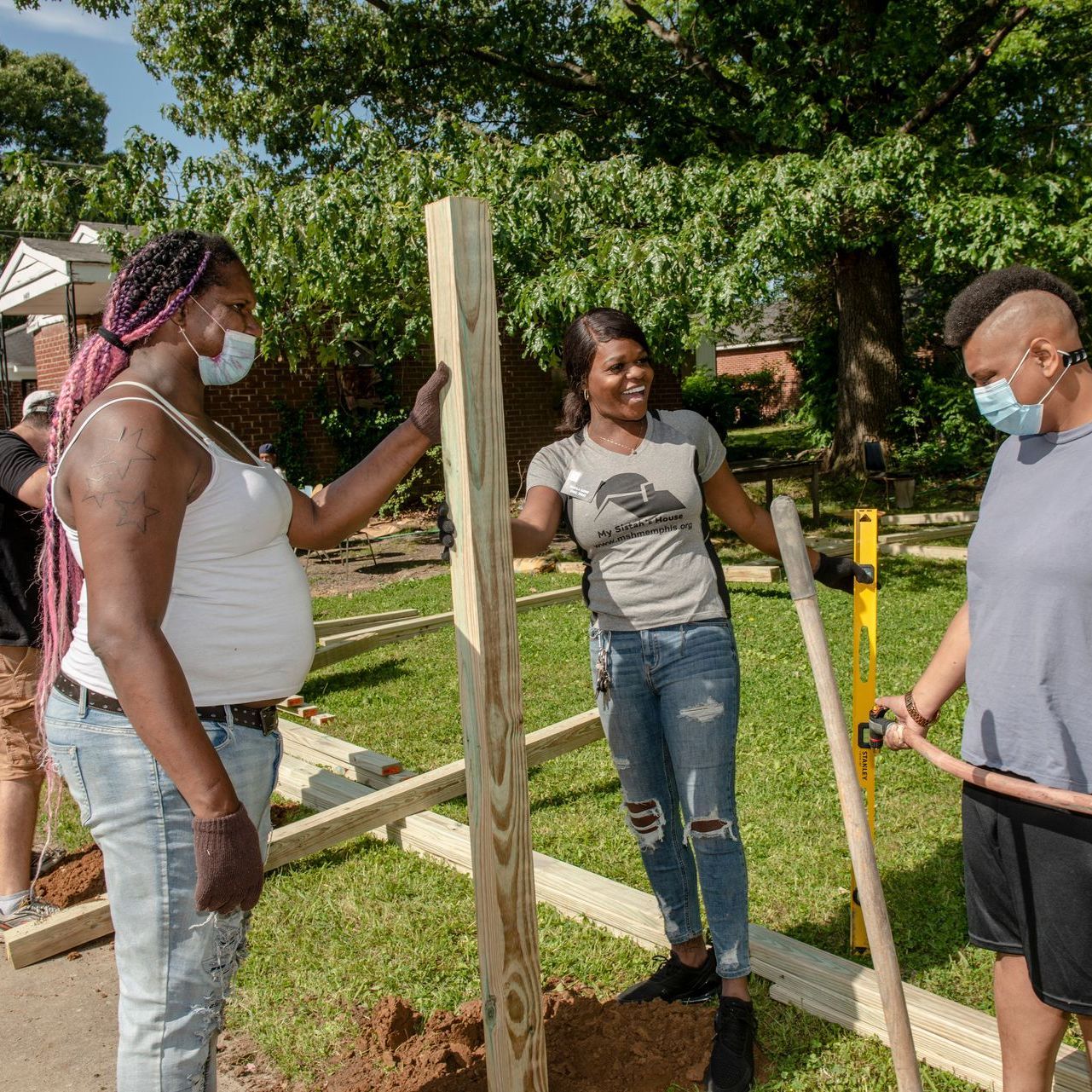 Three masked people assembling a wooden fence post in a sunny yard