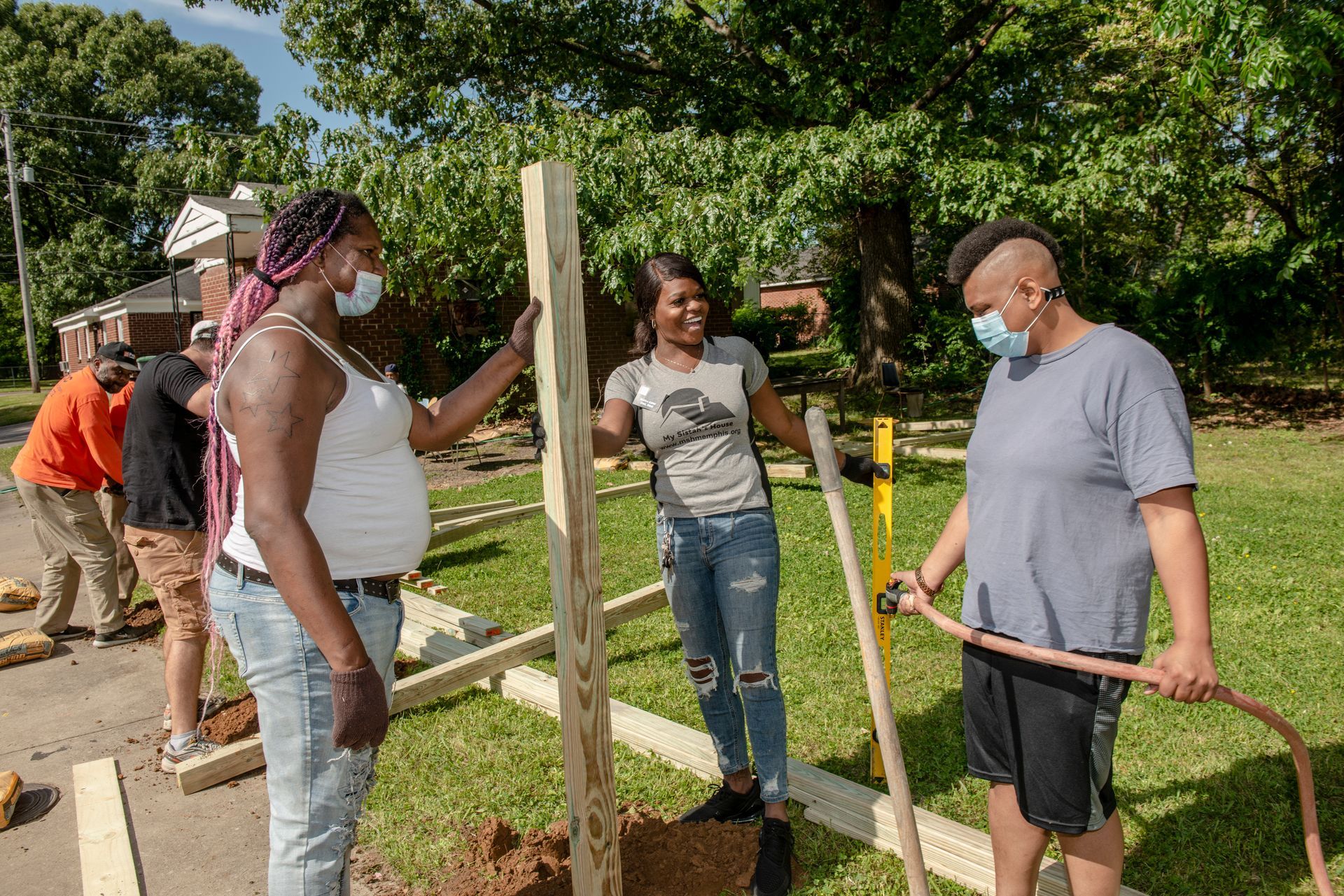 People building a wooden structure outdoors, with tools and a wheelbarrow in a sunny yard.