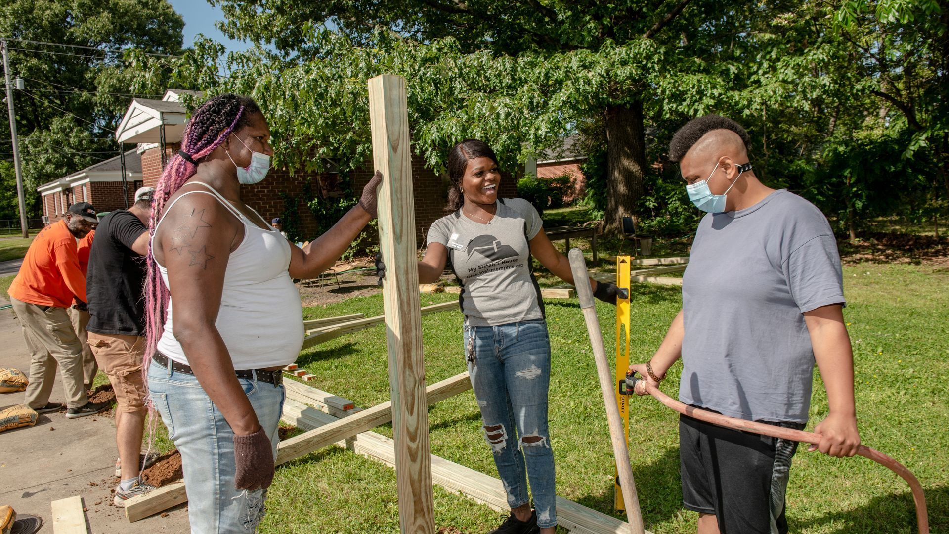 Neighbors planting a tree in a sunny yard, working together with shovels and gloves.