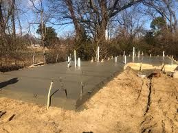 Freshly poured concrete foundation with rebar and a dirt trench beside bare trees under a clear sky