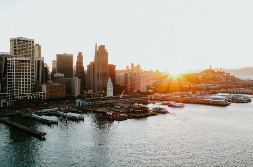City skyline at sunset over a bay; sun flares, buildings and piers silhouetted, soft light.