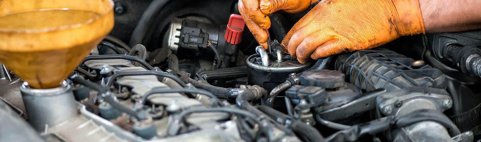 Close-up of hands in orange work gloves performing maintenance on a vehicle engine with a funnel inserted.