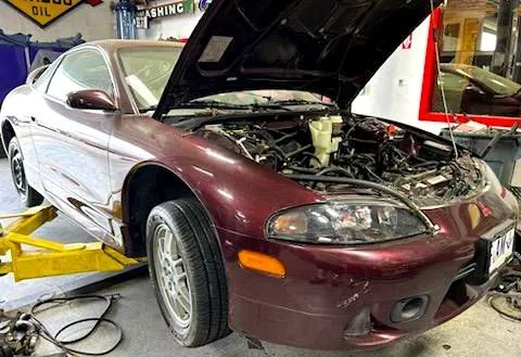 A red car with the hood up is on a lift in a garage.