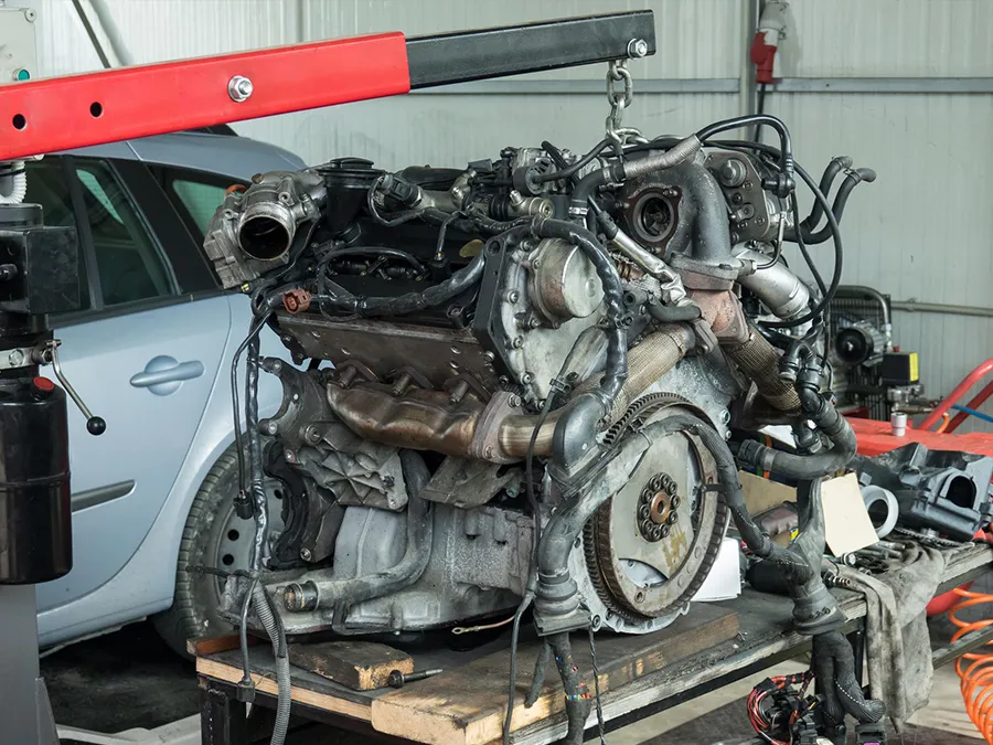 Engine being lifted by a red engine hoist; a car in the background, mechanic's workspace.