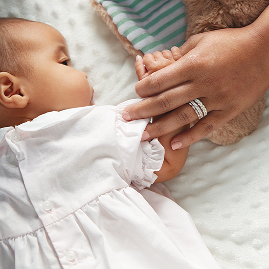A baby in a white dress is being held by a person with a ring on their finger