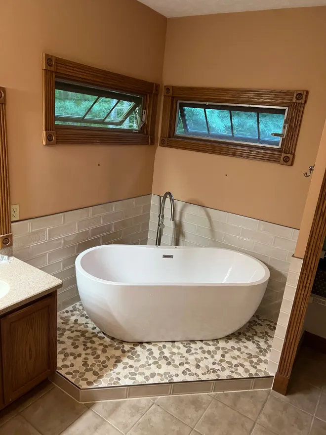 A white, freestanding oval bathtub in a tiled corner with two small, wood-framed windows above it.