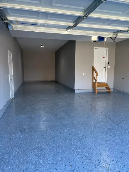 An empty garage with a grey speckled epoxy floor, light grey walls, and a white door with wooden stairs.
