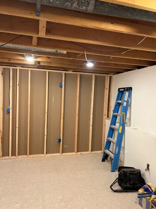 An unfinished basement room with wood stud wall framing, electrical boxes, exposed ceiling joists, a ladder, and tool bag.
