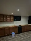 A kitchen under construction featuring brown lower cabinets, one set of wall cabinets, a black appliance, and a bucket.