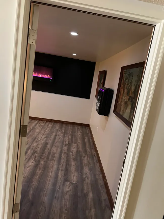 A view through a doorway into a room with gray wood-look flooring, a black accent wall with a fireplace, and wall art.
