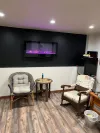 A room features two chairs and a small table arranged under a black wall with a recessed, glowing purple electric fireplace.