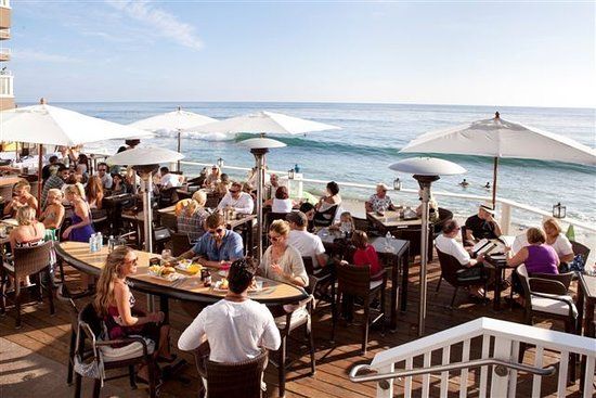 A group of people are sitting at tables on the deck in Laguna Beach overlooking the ocean.