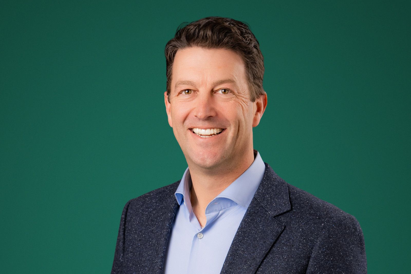 Executive headshots Newport Beach ca Man in blue button-down shirt and patterned jacket smiling against a green background.